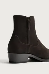 Bota estilo cowboy de caña corta para hombre, confeccionada en piel con acabado serraje de color marrón oscuro. Presenta diseño liso con punta ligeramente cuadrada y tacón bajo, incorporando costuras decorativas laterales.