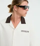 Camisa de manga corta, corte boxy, color crema, con cuello inglés y botones frontales. Presenta un bordado pequeño en el pecho izquierdo con texto en color marrón oscuro.