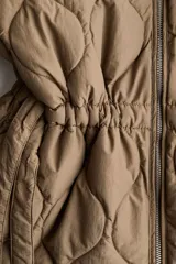 Campera acolchada marrón tipo puffer de nylon guateado, con diseño matelasseado de rombos, cuello redondo y cierre frontal. Cuenta con hombros caídos, mangas largas, bolsillos laterales discretos y cordón de ajuste en la cintura con tiras colgantes.