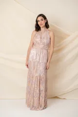 Vestido largo de gasa de seda natural labrada, color marfil, con estampado metalizado en tonos rosados. Presenta un escote halter con abertura en el pecho y una cintura marcada con fajín de strass. La falda es larga y con volados en capas.