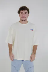 Remera color crema de corte holgado, con cuello redondo y estampado pequeño en el pecho con la frase 'Karma Club' en azul y rojo.