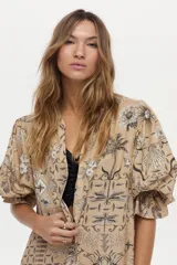 Vestido túnica de lino y viscosa, color camel con estampa artesanal en tonos oscuros, escote en V profundo, mangas amplias abullonadas y falda larga con volado en el ruedo.