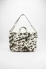 Bolso tipo tote bag con estampado animal print de vaca en blanco, negro y marrón. Cuenta con dos asas de mano y una correa larga para llevar al hombro.