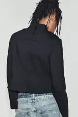 Blazer corto de color negro con cuello mao, manga larga y cierre frontal con cremallera. Presenta doble botonadura decorativa con botones dorados y forro interior a tono.