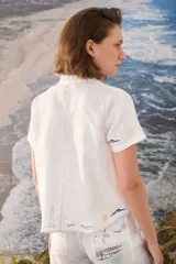 Blusa blanca de lino con mangas cortas, cuello alto con volado y cierre lateral cruzado con botones dorados. Presenta detalles bordados artesanales a contratono en el ruedo y las mangas.