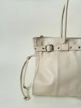 Bolso grande de cuero marrón con textura tipo cocodrilo, estructura rígida y asas para llevar al hombro. Tiene una tira con tachas y hebillas en cada extremo, también lleva lazos en los costados.