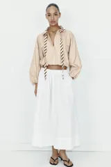 Camisa de popelín color beige con cuello solapa y lazada frontal en contraste con estampado de rayas marrones. Presenta mangas largas abullonadas con puños elásticos, bolsillos con solapa en el pecho y bajo elástico.