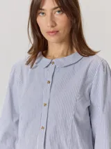 Camisa de denim con silueta levemente entallada, cuello bebé y mangas largas con sutil frunce en los hombros. Presenta costuras curvas decorativas con pespuntes en contraste y cierre frontal con botones.