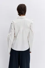 Camisa de color azul oscuro, con cuello clásico, cierre frontal de botones y detalle de volados en los hombros y a lo largo de las mangas largas.