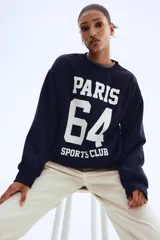 Buzo azul marino de corte oversized con cuello redondo ribeteado acanalado y diseño de texto blanco estampado en el frente que dice 'Paris 64 Sports Club'. Modelo con hombros caídos y remate acanalado en puños y bajo. Interior cepillado suave.