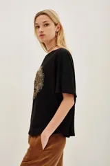 Remera negra de corte holgado con cuello redondo y mangas cortas. Presenta un aplique central en forma de corazón bordado con hilos y cuentas doradas, con un diseño detallado de aves en su interior.
