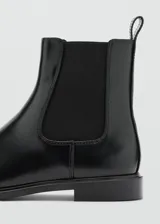 Botín tipo Chelsea de cuero vacuno color negro, con paneles laterales elásticos y tirador trasero. Presenta una silueta clásica con punta redondeada y suela plana de resina de poliuretano.