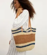 Bolso tipo tote de paja tejida con diseño de rayas horizontales en color natural, marrón y azul marino. Posee asas de hombro en cuero sintético color camel y un dije metálico circular en el frente.