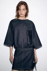 Vestido corto de denim azul oscuro, con cuello redondo, mangas amplias de largo tres cuartos y cintura elástica con cinturón integrado del mismo tejido. Presenta costuras a contraste y cierre posterior con cremallera oculta.