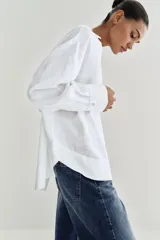 Blusa blanca confeccionada en lino, con cuello polo, escote en V y mangas largas amplias. Presenta un diseño holgado y bajo asimétrico.