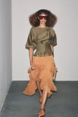 Blusa de manga corta con escote barco, confeccionada en tejido satinado color verde oliva. Presenta un diseño holgado en la parte superior y un canesú inferior con pliegues verticales ajustados.