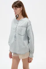 Blusa de jean ligera en color celeste claro, con cuello redondo y cierre frontal parcial con botones. Presenta un diseño con volados a lo largo de la botonera, bolsillo ojal en el pecho, y mangas largas abullonadas con puños ajustados y fruncidos.