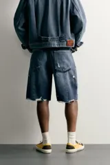 Bermuda baggy fit confeccionada en denim de algodón con lavado azul y efecto rotos en toda la prenda. Presenta diseño de cinco bolsillos, detalles de costuras en las perneras, bajo acabado irregular y cierre frontal con cremallera y botón.