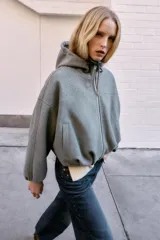 Chaqueta estilo bomber de corte oversize en color marrón oscuro, confeccionada en tejido suave. Presenta capucha con cordones ajustables, cierre frontal con cremallera, mangas largas con puños elásticos y bolsillos delanteros de vivo. El bajo cuenta con detalle de pliegues.