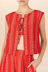 Chaleco sin mangas de color rojo con estampado de cadenas doradas en disposición vertical. Presenta escote redondo y cierre frontal mediante cordones entrelazados.