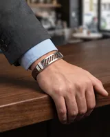 Pulsera compuesta por una banda de acero inoxidable con diseño calado geométrico y una correa de cuerina trenzada en color marrón.