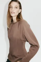 Buzo de felpa con interior cepillado, cuello redondo acanalado y mangas largas raglán. Presenta costuras moldeadoras en la cintura que crean una silueta entallada con efecto peplum.