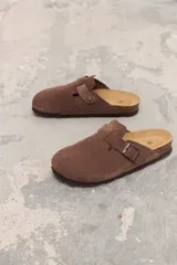 Zueco de gamuza color marrón con diseño de punta cerrada, hebilla metálica lateral ajustable y plantilla de corcho.