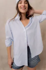 Camisa blanca de calce amplio, confeccionada en tejido liviano con sutiles rayas verticales, cuello clásico, mangas largas con hombros caídos y abotonadura frontal.