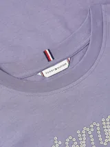 Remera de manga corta, cuello redondo, color lila/malva, con el logo "Tommy" escrito en cursiva con apliques de strass o pedrería. Presenta un pequeño logo de bandera de Tommy Hilfiger en la manga izquierda.