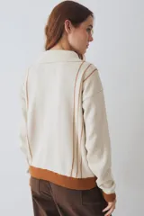 Buzo de tejido de punto color marrón con cuello alto y cierre frontal parcial. Presenta rayas verticales finas en color claro, puños y ruedo en tejido acanalado color crema, y detalles en color verde lima en el tirador del cierre, el cordón de ajuste del ruedo y el parche bordado en el pecho.