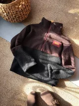 Buzo de lana uruguaya con diseño patchwork geométrico inspirado en el Tetris. Presenta una combinación de texturas y colores en tonos marrón, negro, rojo y gris, con una etiqueta de cuero en la parte inferior trasera.