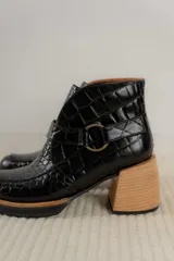 Bota corta de cuero vacuno con textura de reptil, color marrón oscuro. Cuenta con un taco cuadrado negro, suela de plataforma y una tira decorativa con hebilla metálica circular en el tobillo.