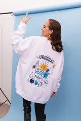 Buzo oversize de cuello redondo en color blanco, con estampa gráfica en la espalda que incluye motivos uruguayos, el sol, la bandera, un jugador de fútbol y elementos tradicionales.
