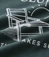 Remera de manga corta y cuello redondo, color verde oscuro, con estampa frontal en blanco que incluye el texto "The Resort Club", una silla de playa y un sol, con la frase "Where time takes sun".