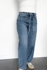 Pantalón de jean estilo mom fit en tono azul clásico, con diseño de tiro alto y lazo ajustable en la cintura. Presenta un corte relajado y holgado en las piernas, confeccionado en denim rígido con estructura.
