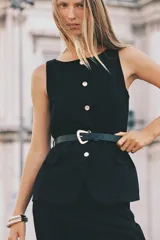 Chaleco negro de vestir, sin mangas, con cuello redondo y cierre frontal de botones metálicos dorados. Incluye dos bolsillos frontales con solapa y un cinturón negro a tono con hebilla metálica dorada.