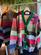 Saco de lana tejido con diseño de rayas horizontales multicolores, cuello en V con borde acanalado en color mostaza y cierre frontal con botones grandes. Presenta un detalle bordado en relieve en la parte superior delantera y mangas largas.