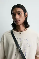 Camiseta de corte holgado y estilo cropped, color beige con efecto lavado. Presenta cuello redondo, mangas cortas y una estampa frontal con texto en azul que simula trazos de rotulador.