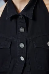 Chaleco de denim negro con escote halter, cuello camisero y cierre frontal con botones metálicos. Presenta dos bolsillos frontales con solapa y diseño sin mangas.