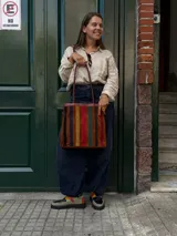 Cartera tipo tote de cuero con diseño de franjas verticales multicolores en tonos tierra, rojo y azul. Cuenta con correas trenzadas de cuero y cierre superior.