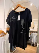 Remera negra de corte relajado, con escote en V, mangas cortas y bajo asimétrico más largo en la espalda. Presenta estampas con escritura estilo manuscrita y un bolsillo decorativo con apliques de strass, encaje y tachas metálicas.