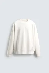Sudadera relaxed fit color blanco crudo, confeccionada en tejido técnico interlock de algodón con elasticidad. Tiene cuello redondo, manga larga y aplicación de etiqueta en el bajo. Los acabados son en rib.