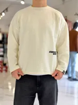 Sweater de punto liviano color marfil con textura marcada, cuello redondo y calce relaxed, con un pequeño bordado negro en el bajo izquierdo.