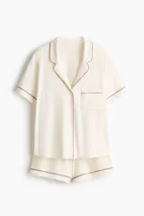 Pijama de dos piezas confeccionado en mezcla de viscosa, compuesto por una camisa de manga corta con cuello abierto, botones frontales y bolsillo superior, y un short con elástico en la cintura. Ambos presentan ribetes decorativos en contraste.