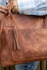 Cartera tipo tote de cuero marrón con costuras artesanales visibles, bolsillo frontal con el logo grabado, asas de hombro y detalles de flecos colgantes.