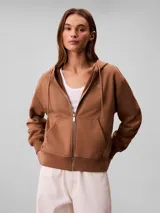 Campera canguro de felpa color marrón, con capucha ajustable con cordones, cierre frontal completo y bolsillos tipo canguro. Presenta el logo retro de Calvin Klein bordado en el pecho.