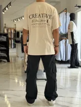 Remera oversize color beige, con diseño estampado en la espalda que incluye tipografía grande y texto descriptivo en negro.