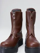 Botas de cuero color marrón de caña alta, con plataforma gruesa de goma y cierre metálico frontal con tirador. Presentan costuras a contratono y detalle de etiqueta de la marca en la lengüeta.