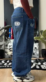 Pantalón de jean azul oscuro de corte wide leg, con diseño holgado y tiro medio. Presenta un pequeño bordado con alas en el bolsillo delantero.