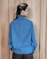 Camisa de denim con volados verticales a lo largo de la botonadura frontal y cuello alto. Presenta mangas largas con puños elásticos y detalle de volados en los mismos.
